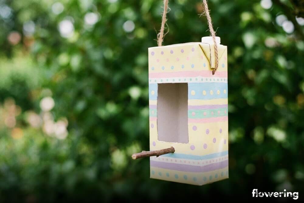 Kolorowy karmnik z kartonu po mleku typu Tetra Pak z okienkiem i żerdką, zawieszony na sznurku