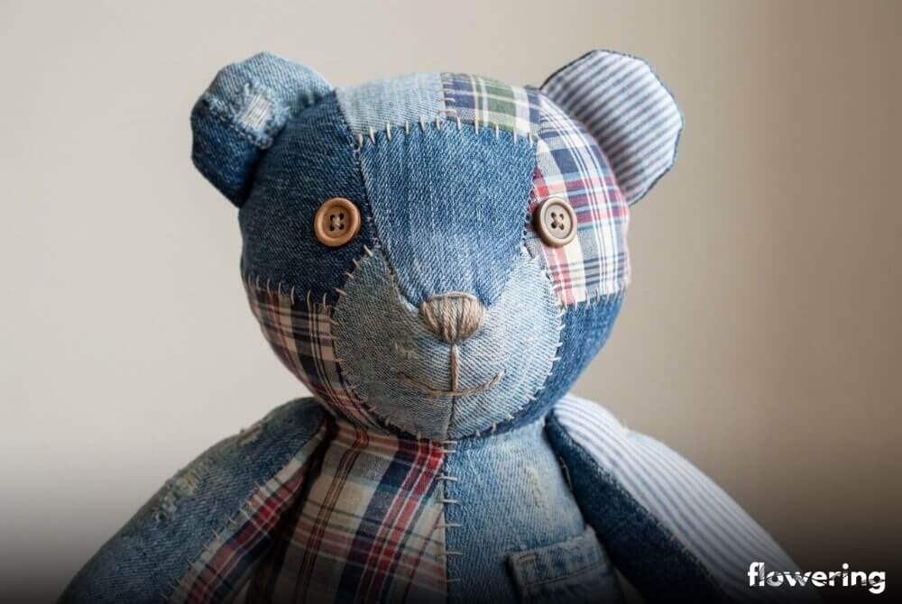 Miś uszyty z fragmentów starych ubrań, z guzikowymi oczami i haftowanym noskiem, pokazany w zbliżeniu na neutralnym tle