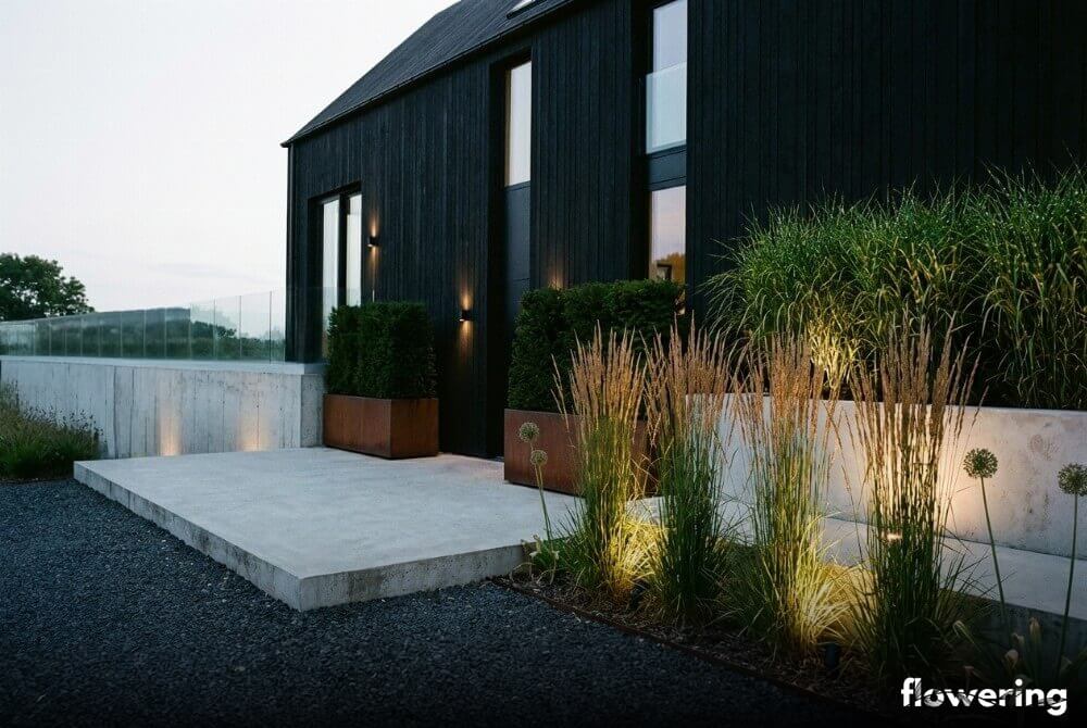 Industrialny ogród przy domu typu stodoła z betonem, stalą kortenowską i ciemnym grysem, z trawami ozdobnymi i dyskretnym podświetleniem punktowym