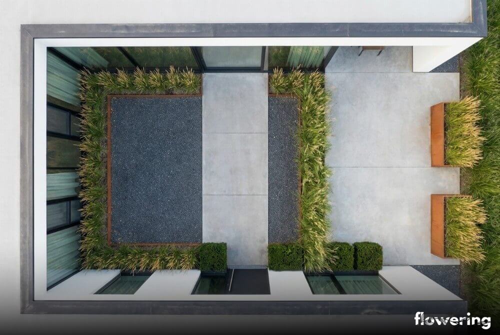 Widok z góry industrialnego ogrodu przy domu z betonem i ciemnym grysem, donicami z kortenu oraz powtarzalnymi nasadzeniami trzcinnika, miskanta i cisa